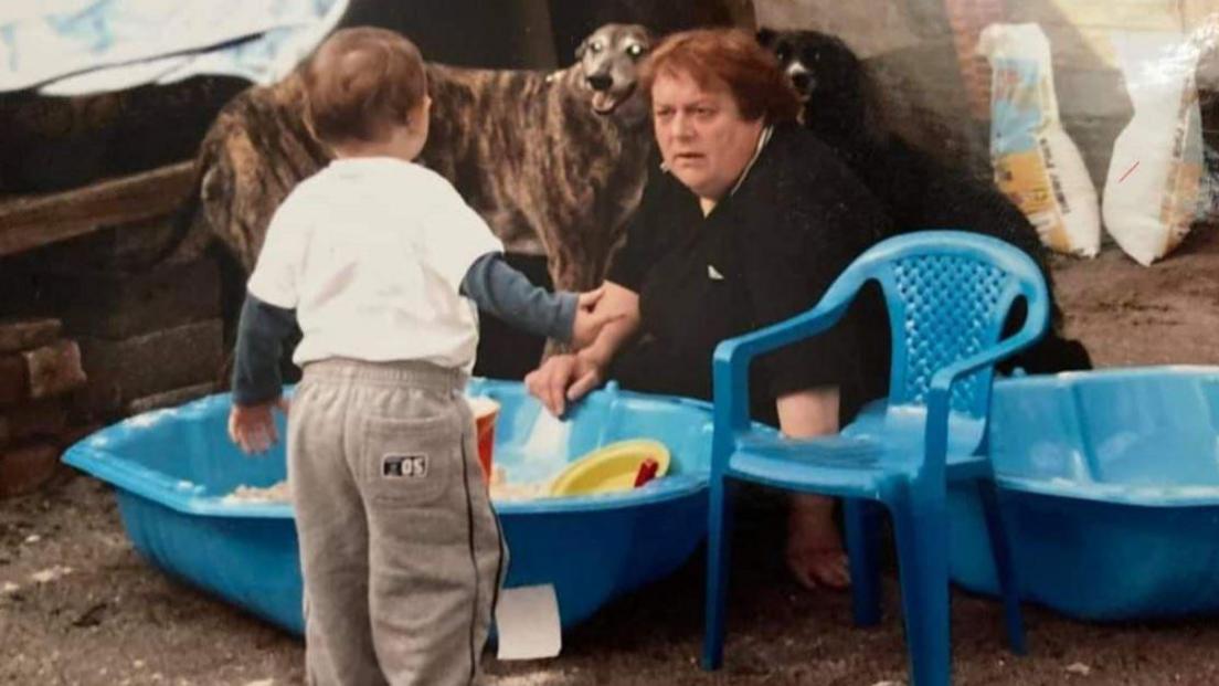 A dog in the background as a little boy looks at a middle-aged woman sat between children's toys