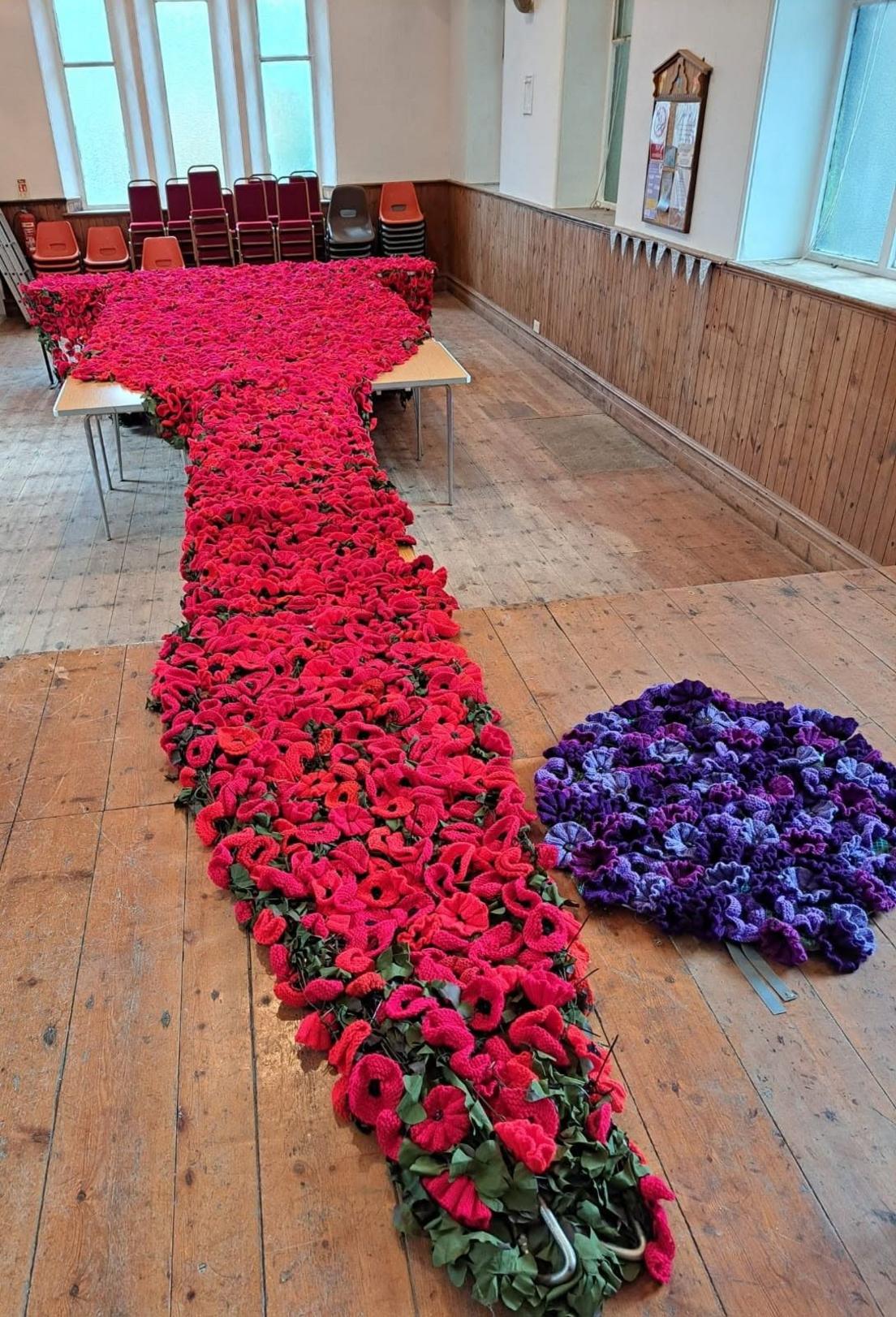 The red poppy display is seen rolled out on a wooden floor. To the side of it is a smaller purple knitwork.