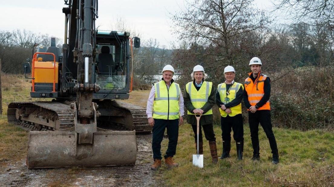 Four men, including Lord Bath, wearing hi-vis vests and hard construction helmets. Lord Bath is holding the handle of a spade, and they are posing for the camera beside a large digger. It is an overcast grey day in winter, the ground is muddy and the trees have been stripped of their leaves. 