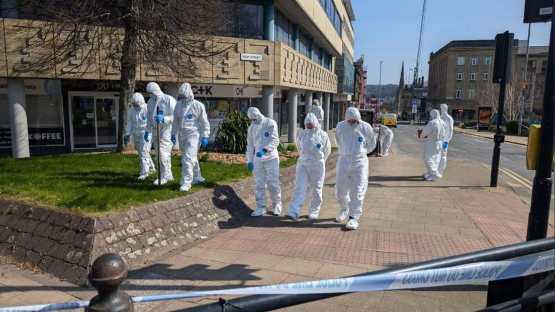 A number of forensic officers in white suits walk through Huddersfield in a crime scene, there is police tape in the foreground.