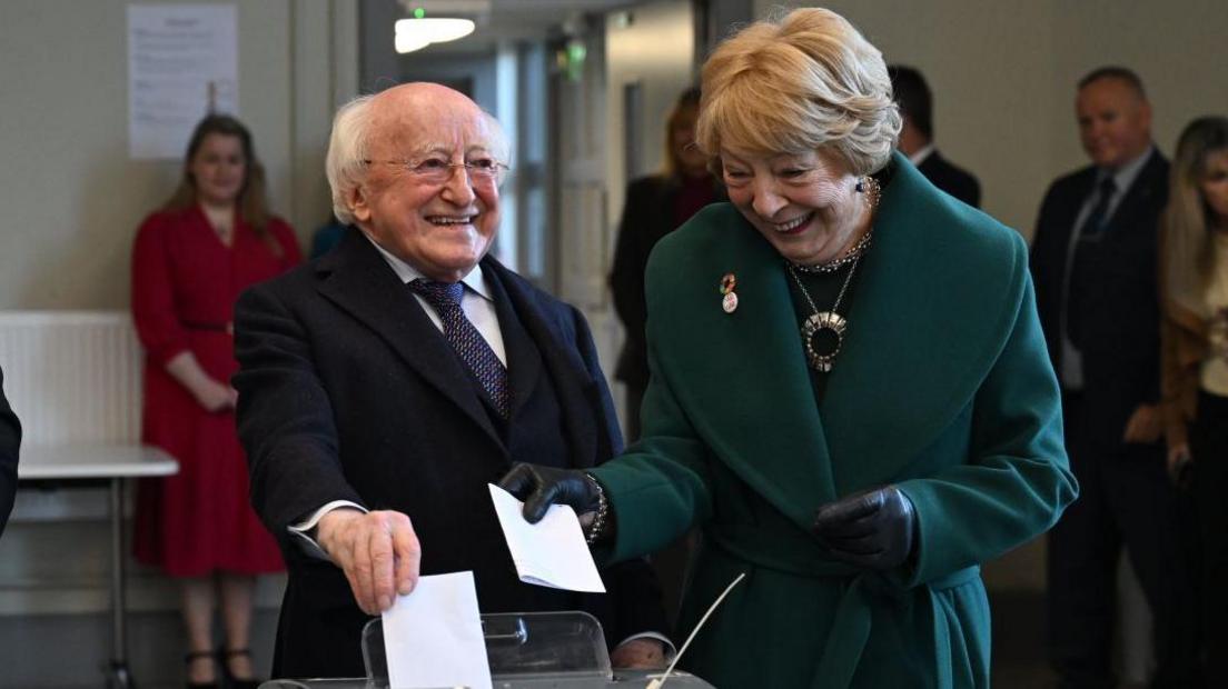 A short man with glasses and white hair stands next to a woman with blonde hair. They are both putting pieces of paper into a box while smiling. The man is wearing a suit, while the woman is wearing a green jacket. To their left is a man watching them, smiling. There are also people in the background.