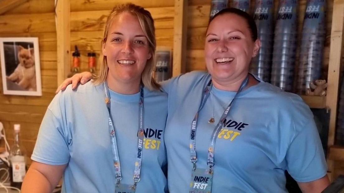 Jody Peters with another woman wearing light blue IndieFest t-shirts while stood in a converted garden shed. Cups and bottles can be seen in the background.