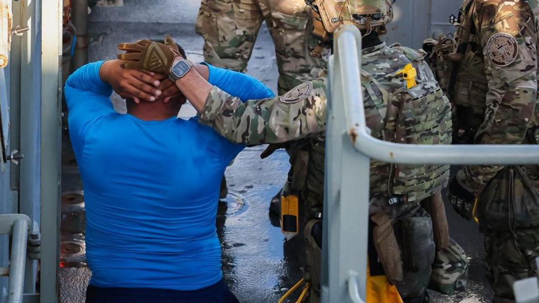 Rear view of a man wearing a blue t-shirt with his hands on his head. His hands are being held in place by a person in a camouflage military uniform