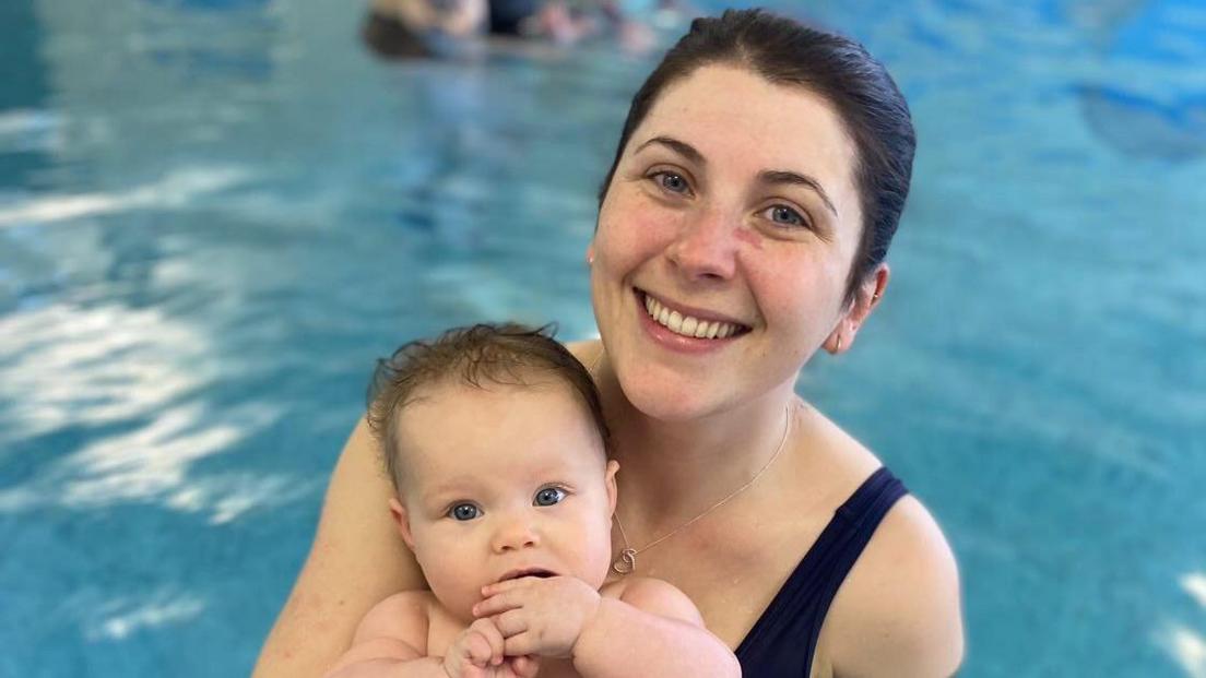 Amy holding her baby Murphy in a swimming pool. She is smiling at the camera. The baby has her hand in her mouth.