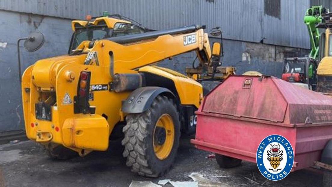 A yellow JCB handler and other plant hire machinery at an industrial estate compound, with a corrugated metal wall in the background. There is a blue and white West Midlands police logo in the bottom right corner.