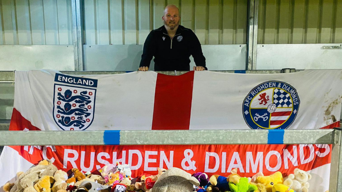 Man standing on a football terrace, with an England flag spread out in front of him, at waist height, with AFC Rushden & Diamonds written in white capitals across the red horizontal stripe. A mound of soft toys can be seen at the bottom of the image.