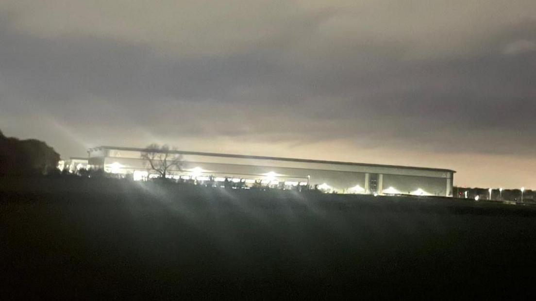 A brightly lit industrial unit, viewed from a field. There are trees between the photographer and the building, and the photograph has been taken at night.