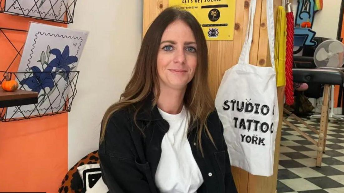 Tattoo studio owner Lisa Harding Mottley is looking straight into the camera. She is wearing a white T-shirt, a dark jacket, and is sitting in her studio.