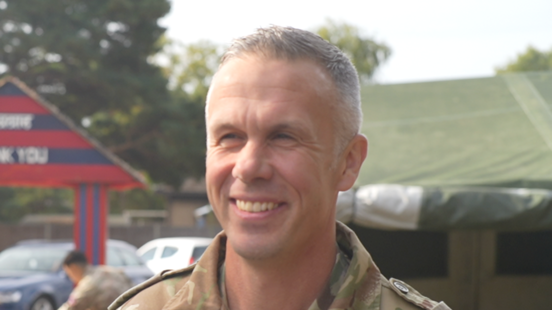 Lt Col David Crosbie smiles as he looks to the left of the camera. He has short grey hair and wears a green Army uniform. Green tents can be seen behind him as well as another soldier. 