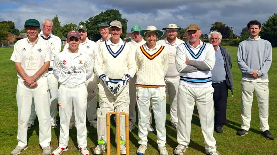 A men's cricket team pose for the camera. They are all wearing cricket whites and are posing for the camera and smiling. Gerald Cooke is in front of the stumps.
