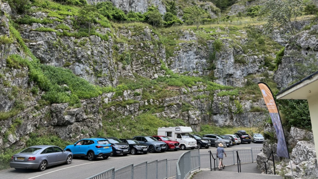 Cars parked along a winding road running through Cheddar Gorge. The road is quite narrow and there is someone walking on the pavement on the right hand side of the road.