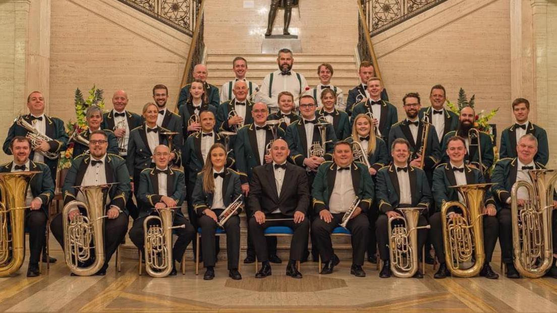 The band is sitting and standing on the steps of a marbled hall in Stormont. There is a mixture of ages among the members - men and women between the age of 15 and 72. They are all wearing white shirts, a black bow-tie and black trousers. Most of the members are wearing a green jacket that has gold embroidered embellishments.