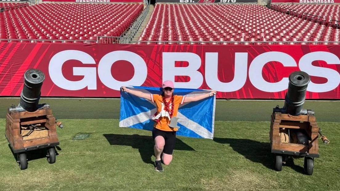 Sarah kneeled down on the field at Raymond James Stadium. She is wearing an orange Buccaneers jersey and is holding a Scotland flag behind her. She is also wearing a large red chain which has the team's logo on. The logo is red with a large white skull and crossed swords on it.