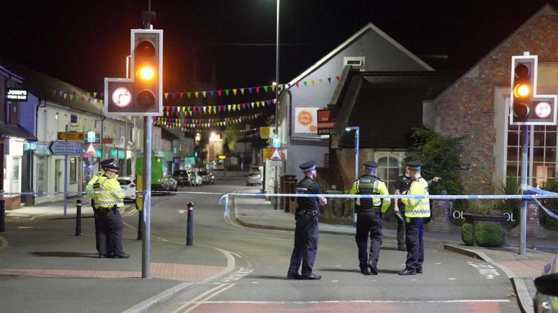 Police standing in the street at the scene. It is dark and a traffic light is on amber. There is no traffic.