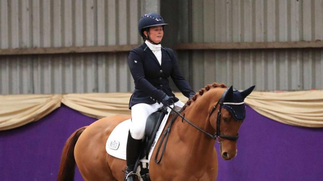 A woman riding a brown horse inside. She is wearing a helmet and a black coat, and boots. 