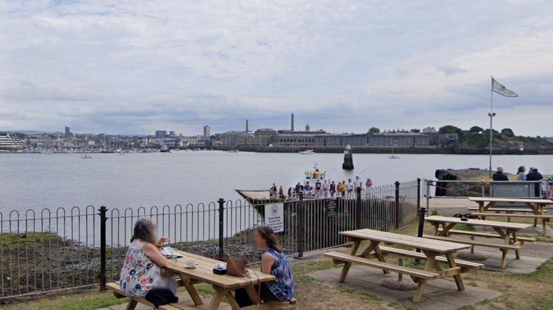 A street view of the Mount Edgcumbe slipway. There are two women sat at a picnic table on the grassy area and there are three other tables behind them. Below you can see people stood on the slipway and the top of the Cremyll ferry which is yellow. It is on the water, which is in the background along with other buildings on the other side of the sea.