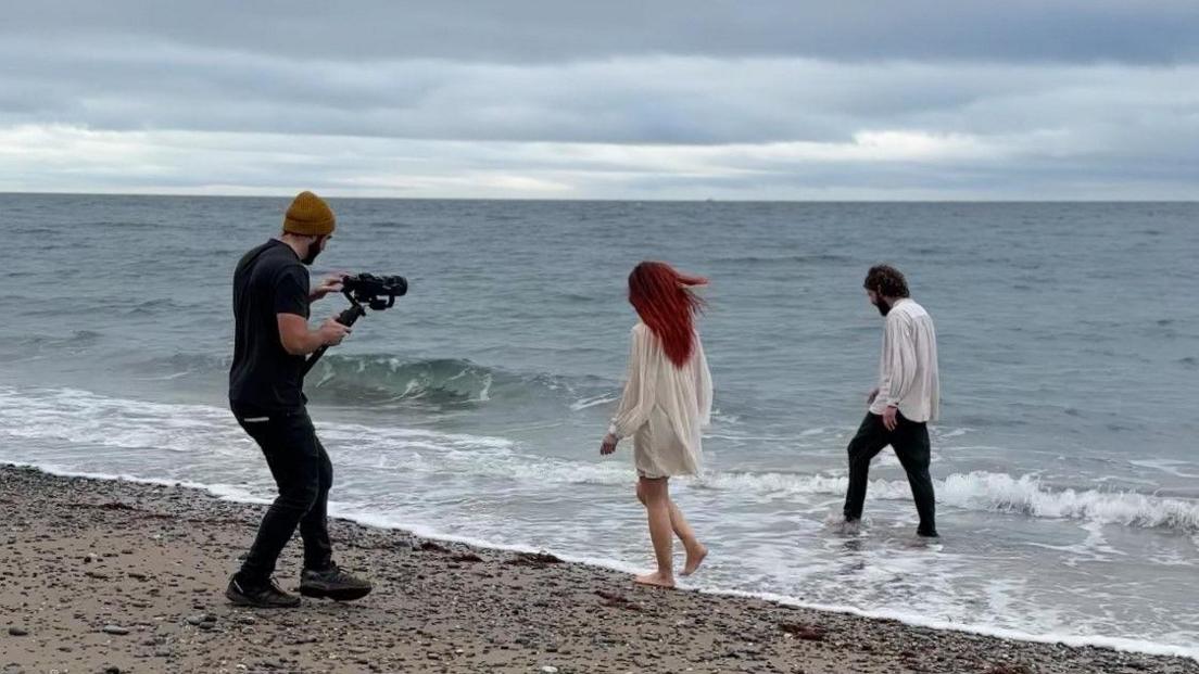 A man with a camera on the left filming a man and a women walking barefoot along the shoreline of a beach on a cloudy day.