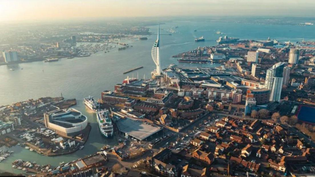 Aerial view of Portsmouth with waterfront around housing and buildings with the Spinnaker Tower on the edge of the city and numerous ships on the water.