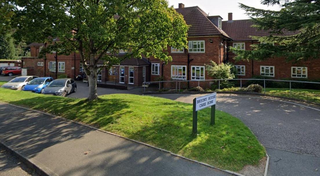A long red brick building viewed from the pavement with carparking and a grass verge in front. A sign on the verge says Bryony House Care Home