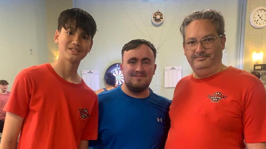 Three men stand together in a hall. The teenager to the left has dark short hair and wears a red t-shirt with a darts logo on the left breast. The man on the right wear a matching t-shirt and wears glasses, with a moustache and grey curly hair. The man stood between them both is famous darts player, Luke Littler, wearing a blue t-shirt with a soft brown beard and short hair.