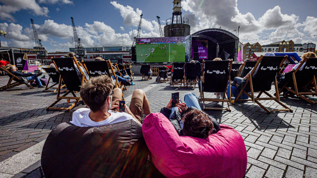 Two people are lying on beanbags, with rows of people sat in chairs in front, all watching a large screen display a rugby match