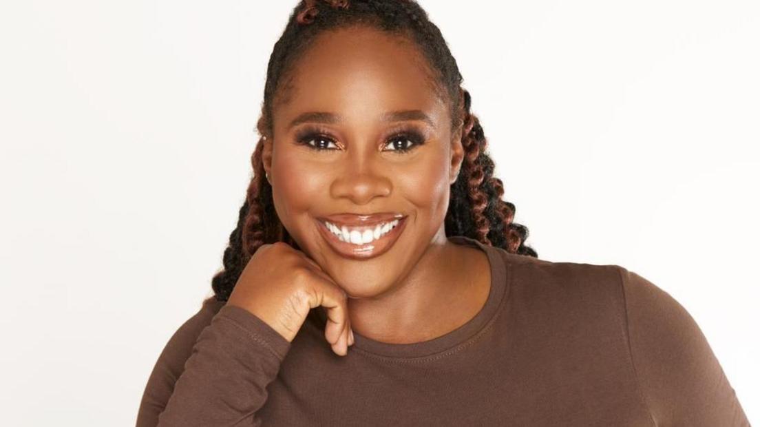 Denise is wearing a brown top and is pictured against a plain white background. She has long, black braids with curls and has her chin resting on her hand and is smiling.