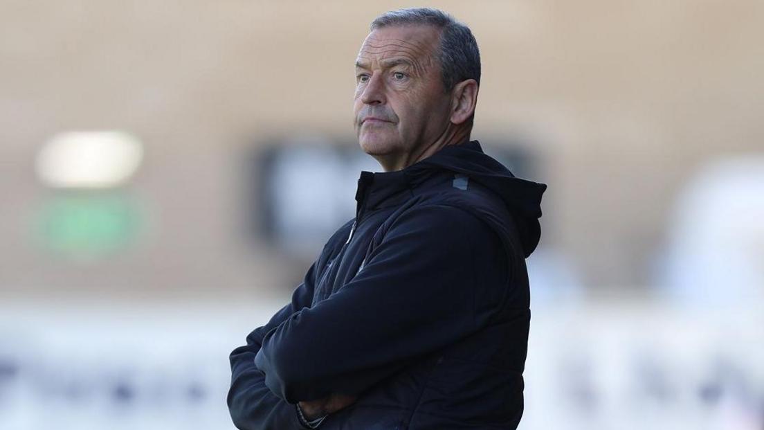 Colin Calderwood is wearing a black jacket with a hood standing with his arms folded watching a game from the touchline.