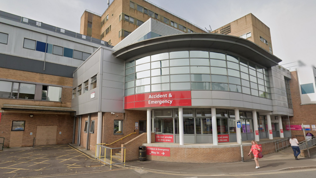 Exterior of Yeovil District Hospital's A&E department. The building is a mixture of brown bricks and grey panels with A&E sign. There are a few people walking around near the entrance. 