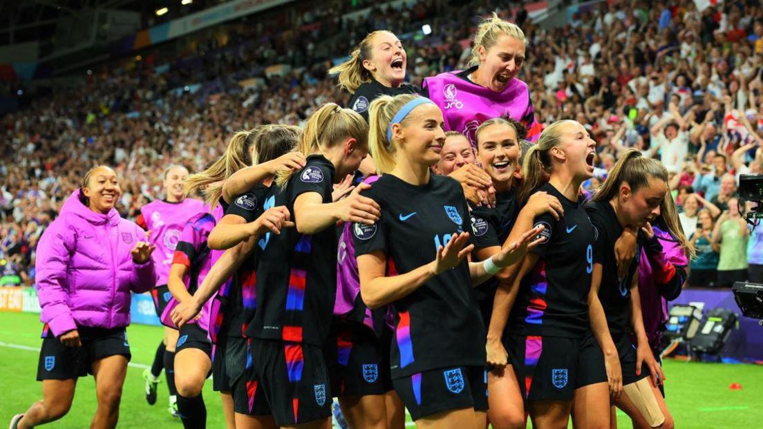 Multiple members of England Women's football team face the crowd as they huddle in a post-goal pitch celebration.