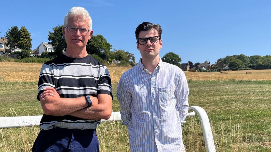 Two men stand in front of a golden brown arable farm field which has stubble where it has just been harvested.
The man on the left has his arms folded and is wearing a black and white striped top. The man to his right has a white shirt with blue vertical stripes.
the are standing in front a rail with a green horse track behind them.