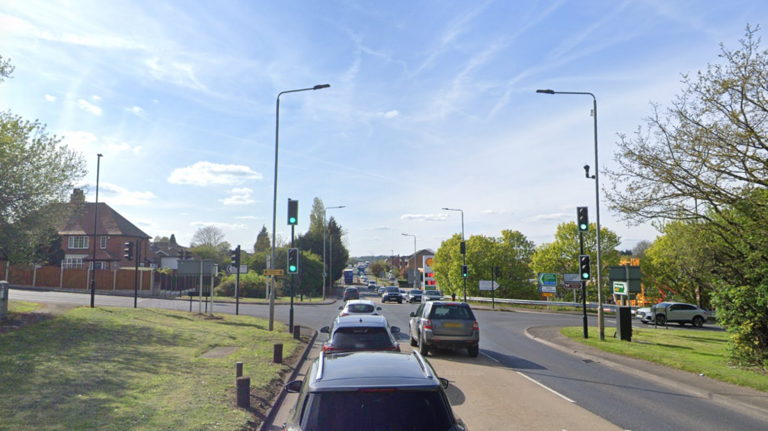 Cars queue at a traffic junction on the A57. The crossroads is surrounded by trees and grass verges and there is a Shell garage in the distance. 