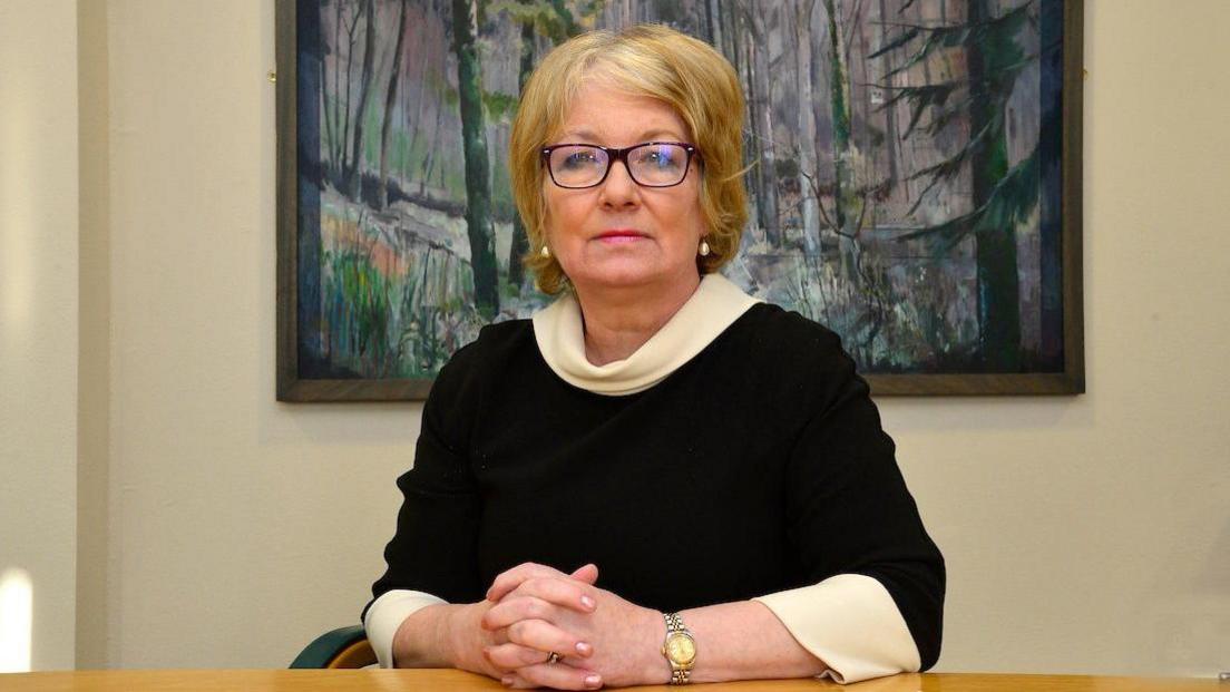 Marie Anderson is a middle-aged woman with blond hair and black-rimmed glasses. She is wearing a black jumper with white sleeves and neck and is at a desk with a a painting of trees in the background