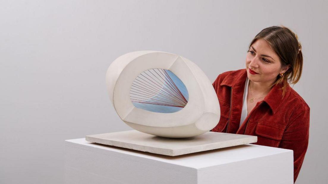 A woman in a red jacket looks at the sculpture in a white room.