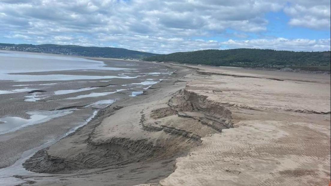 It is a cloudy day over Morecambe Bay with some blue patches of sky. There are patches of water along the bay with steep drops evident in parts of the sand. 