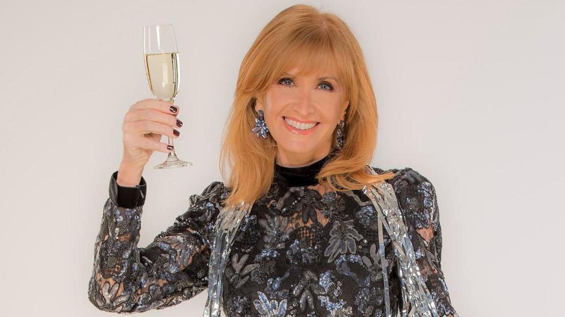 Jackie Bird smiles at the camera while raising a glass of wine. She has strawberry blonde hair and wears a sparkly silver and black long-sleeved top.