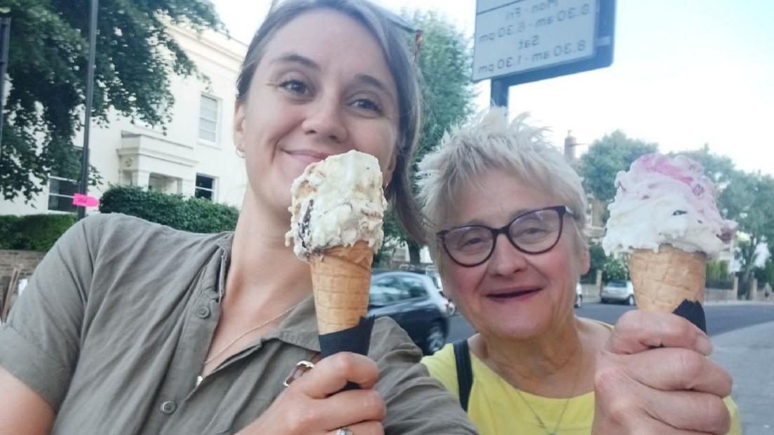 Cat Rubens and Jane Rubens holding ice creams and smiling. Cat is wearing a green button up shirt and sunglasses, while Jane is wearing a bright yellow t-shirt and glasses.