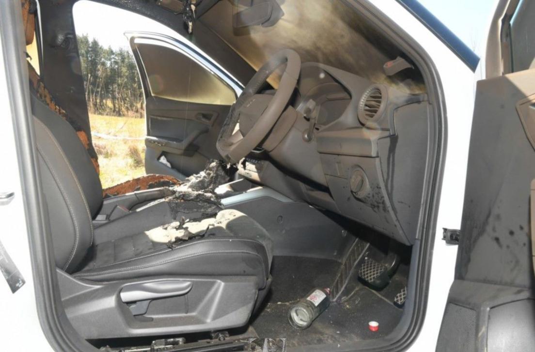 Interior of a white car. It is all covered in soot and the front seats have been burned. An empty glass bottle is in the footwell near the pedals.