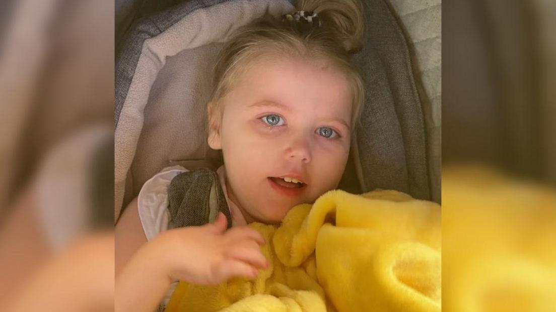 Phoebe sitting in a pram with a bright yellow fluffy blanket. She has blonde hair tied up in a ponytail and light blue eyes.