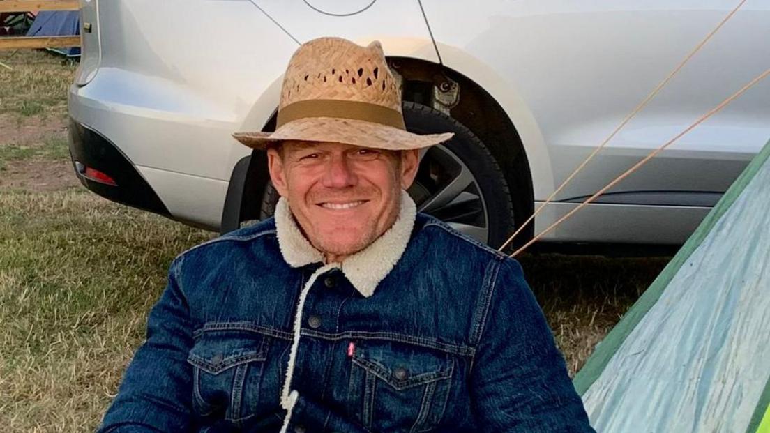 A man sits smiling at the camera. He is wearing a straw hat and demin jacked with a sheeps wool collar. Behind him is the bottom part of a silver car, grass and the edge of a tent. 