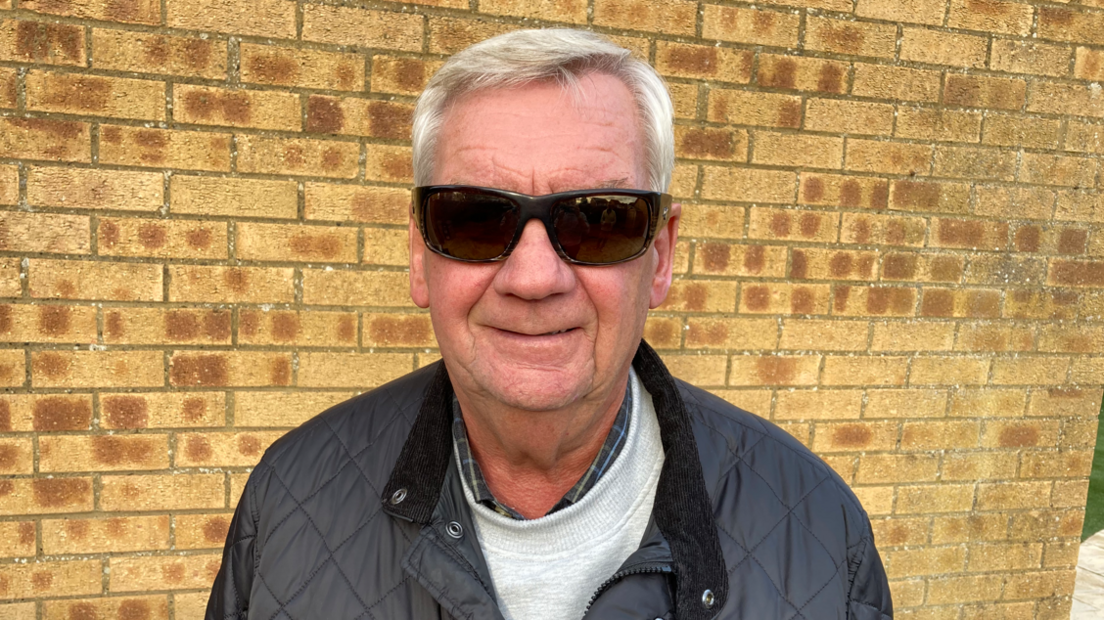 A mid-shot of Alan Fletcher standing in front of a brick wall with sunglasses on