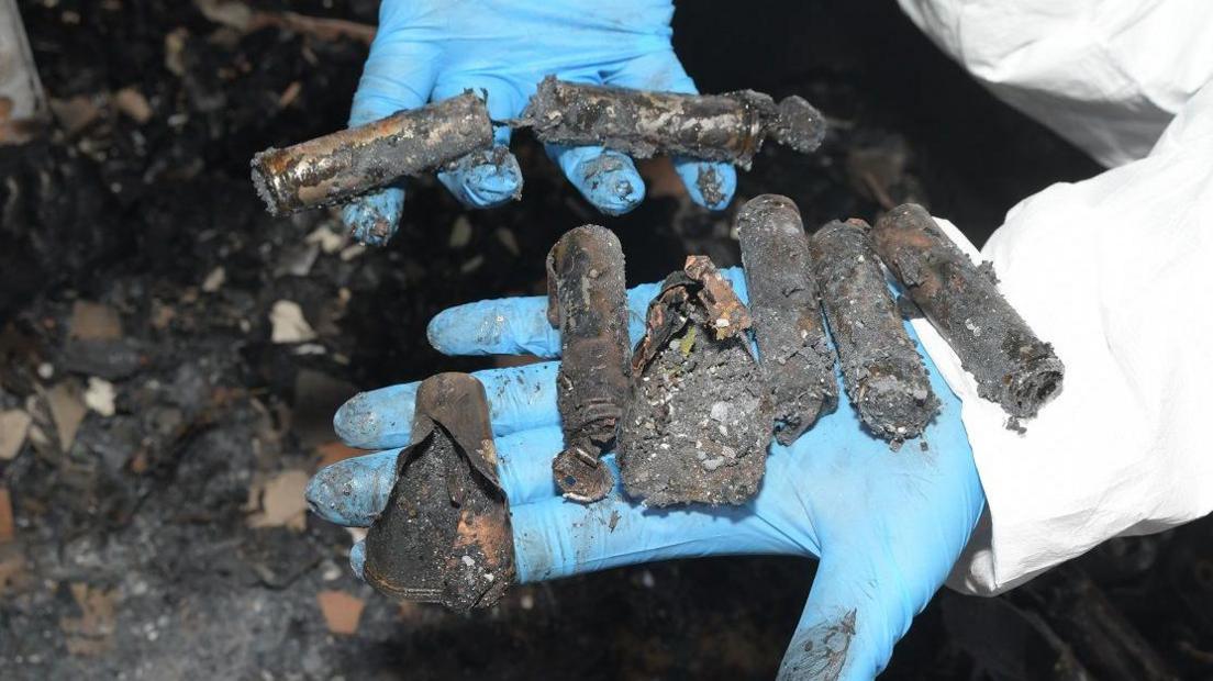 A fire investigator in a white forensic suit and blue plastic gloves holds fragments of an exploded hoverboard in a charred area of a house after a fire.