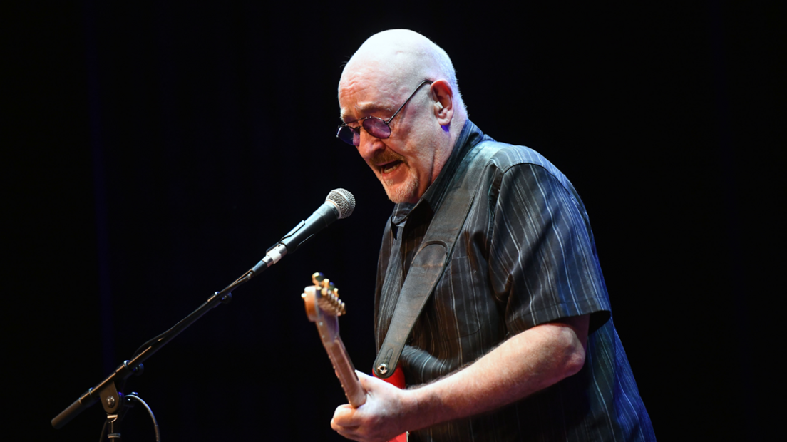 Dave Mason in a dark striped shirt with a red guitar strapped over his shoulder - he plays the guitar and sings into a microphone