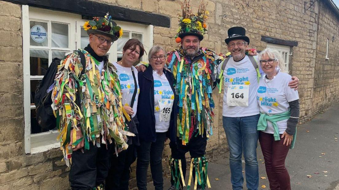 Peter Gale - standing third from left in a group of six people - a man dressed in a Morris coat, a two woman wearing white Sue Ryder T-shirts, Peter in a Morris coat and a man and a woman wearing white Sue Ryder T-shirts. All of them are standing on a pavement by the road.