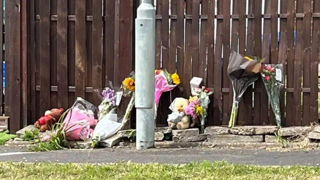 Floral tributes and two teddy bears are laid against a wooden fence by the pavement near the home of Muhammad Esmael