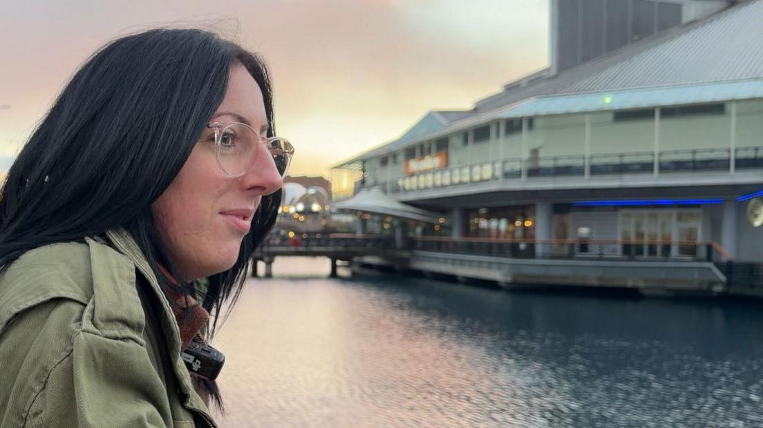 A lady looking across water, the sky is light pink and she has black hair. She's wearing clear glasses and a green coat. 
