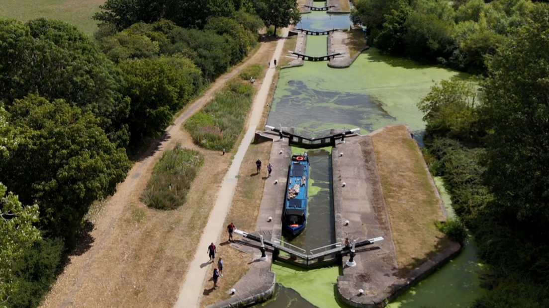 An aerial view of a green canal with a set of locks and trees on either side, with a blue narrowboat between the lock gates