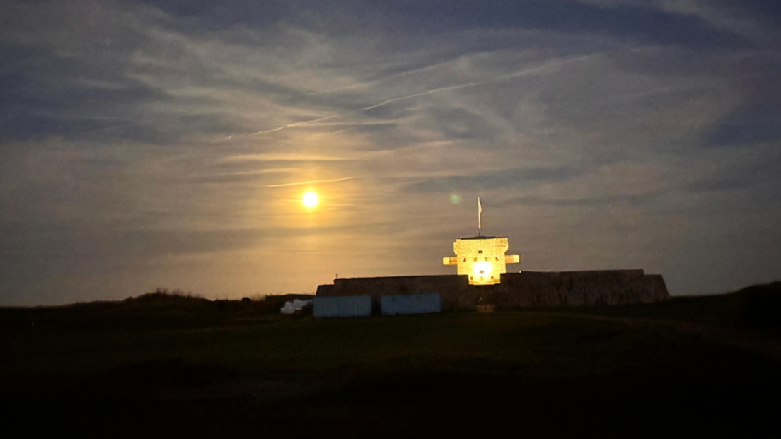 A bright orange moon lighting up the sky. It's slightly restricted by clouds. There is a large building, part of which is lit up, the other in complete darkness. 