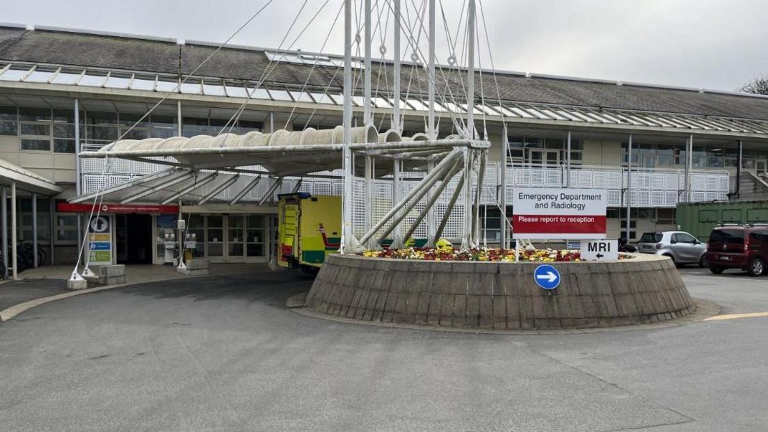 Princess Elizabeth Hospital, La Vauquiedor, St Martin, Guernsey. The image shows the entrance to the Emergency Department and Radiology unit. There is a grey concrete roundabout in front of the entrance with an ambulance parked half out of sight behind the roundabout. The building is grey and cream in colour and comprised of two storeys.