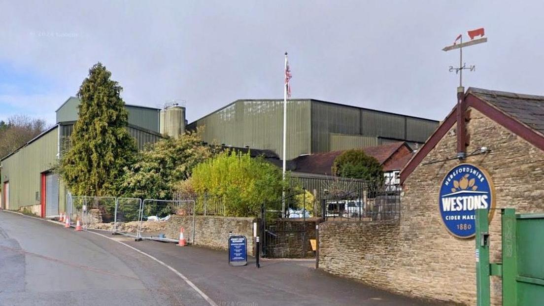 A street view image of the Weston's Cider building which is green. There is a Union Jack flag flying and shrubbery around the building.