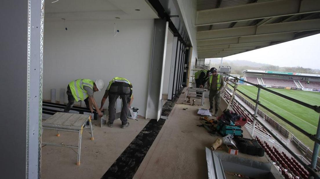 Two men in green high visibility jackets and white hard hats measuring a metal beam standing on the balcony of what will be a box area of the new East Stand at Northampton Town Football Club. 